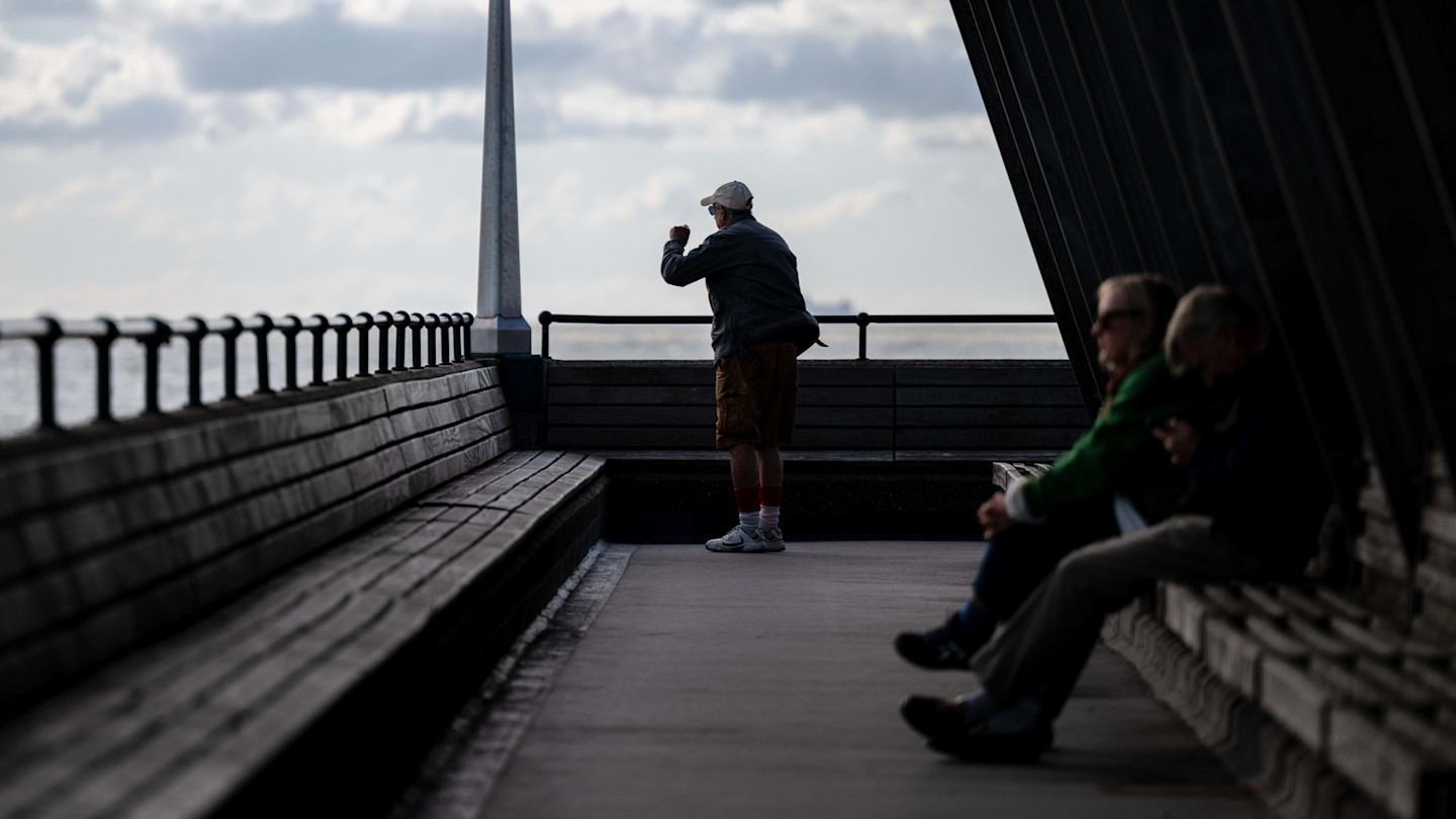An older person stands at the end of a pier looking out to sea, while two people sit on benches in the shade.