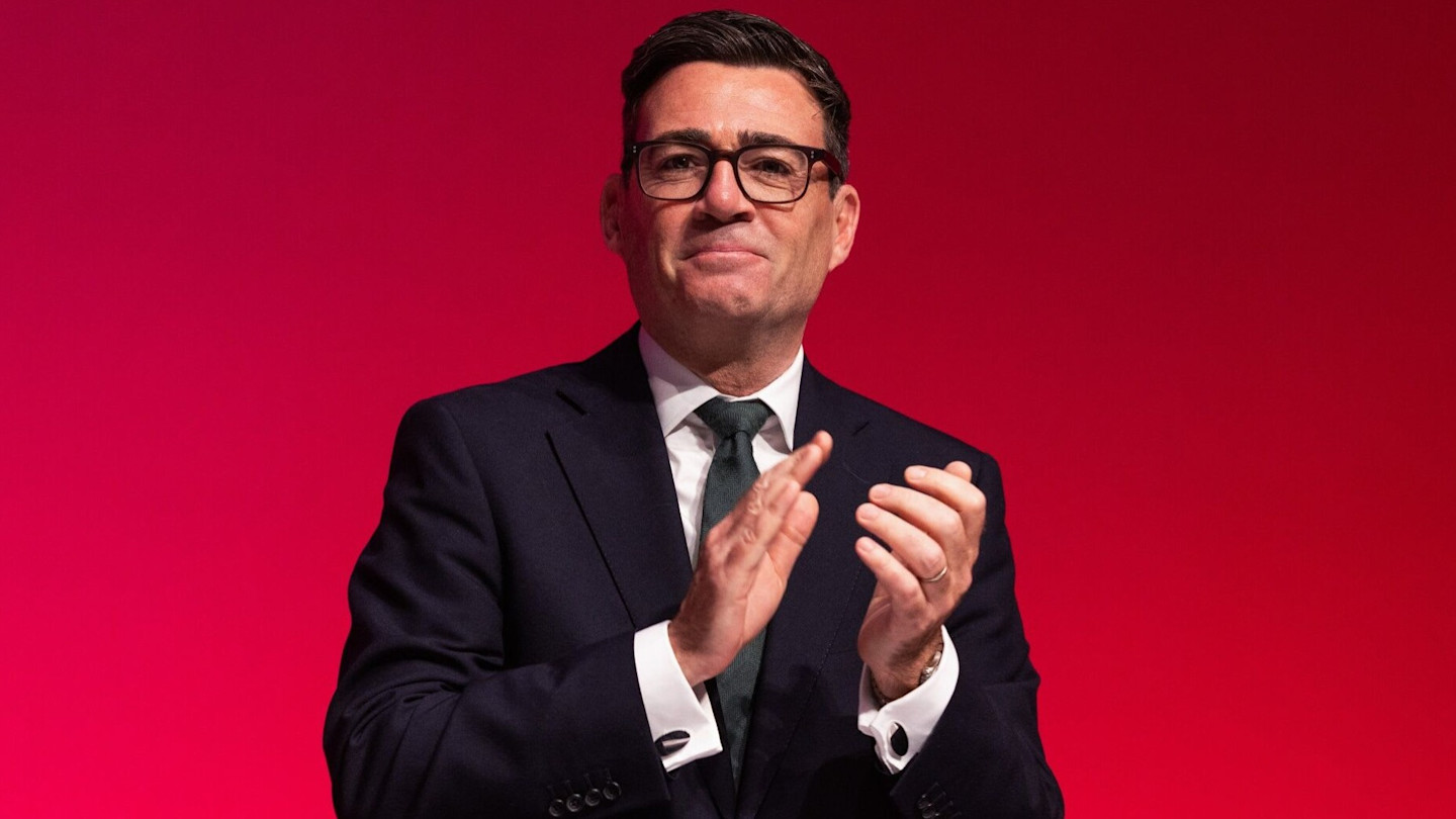Andy Burnham clapping on stage against a red background.