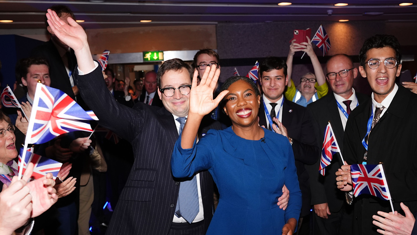 Kemi Badenoch and Hamish Badenoch wave and smile as they walk through a crowd holding Union Jack flags at the Conservative Party conference.
