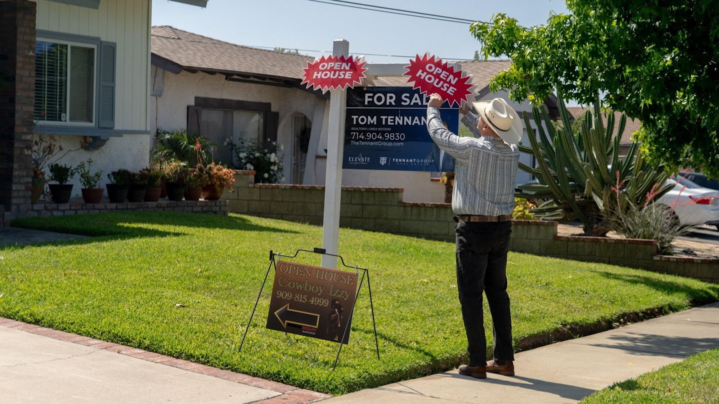 A real estate agent in a cowboy hat sets up "Open House" signs on a "For Sale" post outside a suburban home.