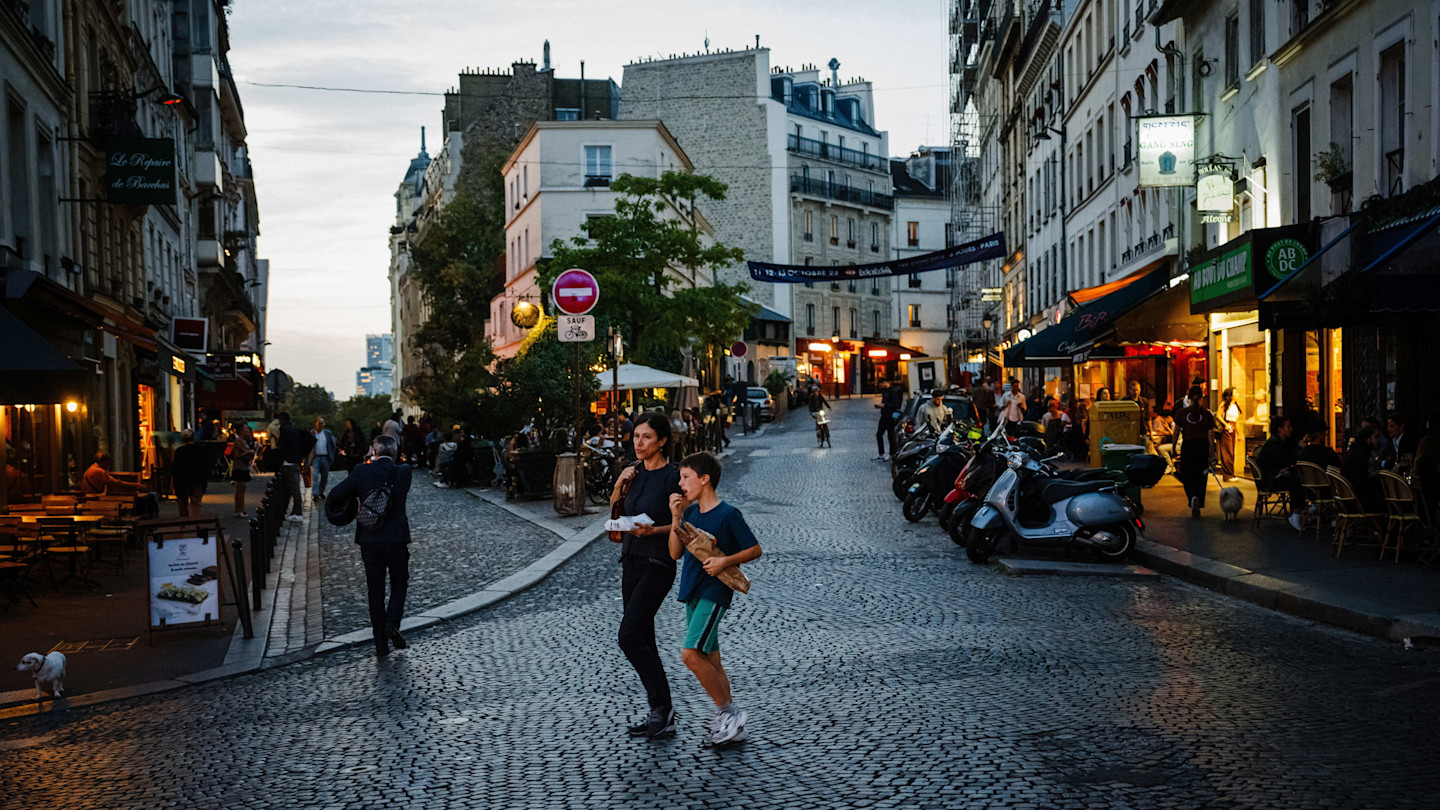 People walk along a cobblestone street lined with cafés and shops in Montmartre at dusk. A woman and child carry baguettes.