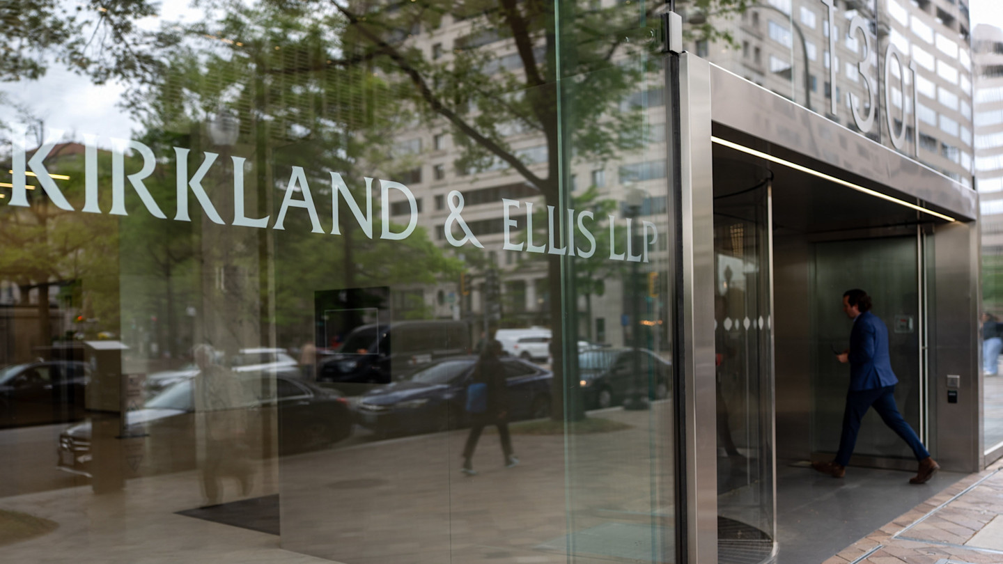 Sign for Kirkland & Ellis LLP is seen on a glass wall at a building entrance, with a person in a blue suit walking inside.