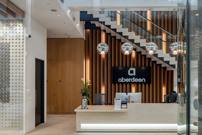 Reception area of the Aberdeen London office featuring a modern desk, three hanging silver globe lights, and the abrdn logo on the wall.