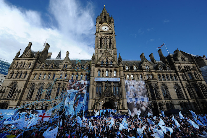 A large crowd of Manchester City fans wave blue flags outside Manchester Town Hall as players and manager Roberto Mancini celebrate their Premier League title on a stage, with banners and streamers decorating the building.