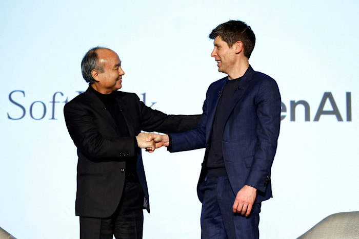 Masayoshi Son and Sam Altman shake hands and smile on stage at an event, with SoftBank and OpenAI logos in the background.