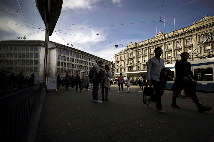 People walk past the headquarters of Credit Suisse and UBS at Paradeplatz in Zurich, with trams and historic buildings visible