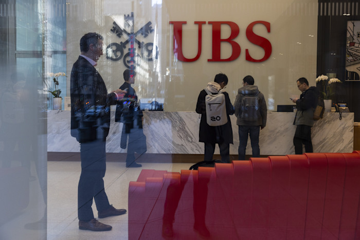 People stand at the marble reception desk under a large UBS logo inside the UBS flagship office, with reflections visible on the glass.