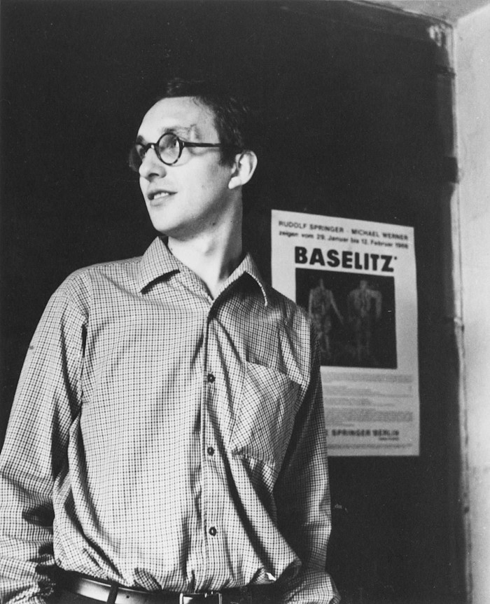 Georg Baselitz stands in front of a “Baselitz” Heroes exhibition poster, looking to the side.