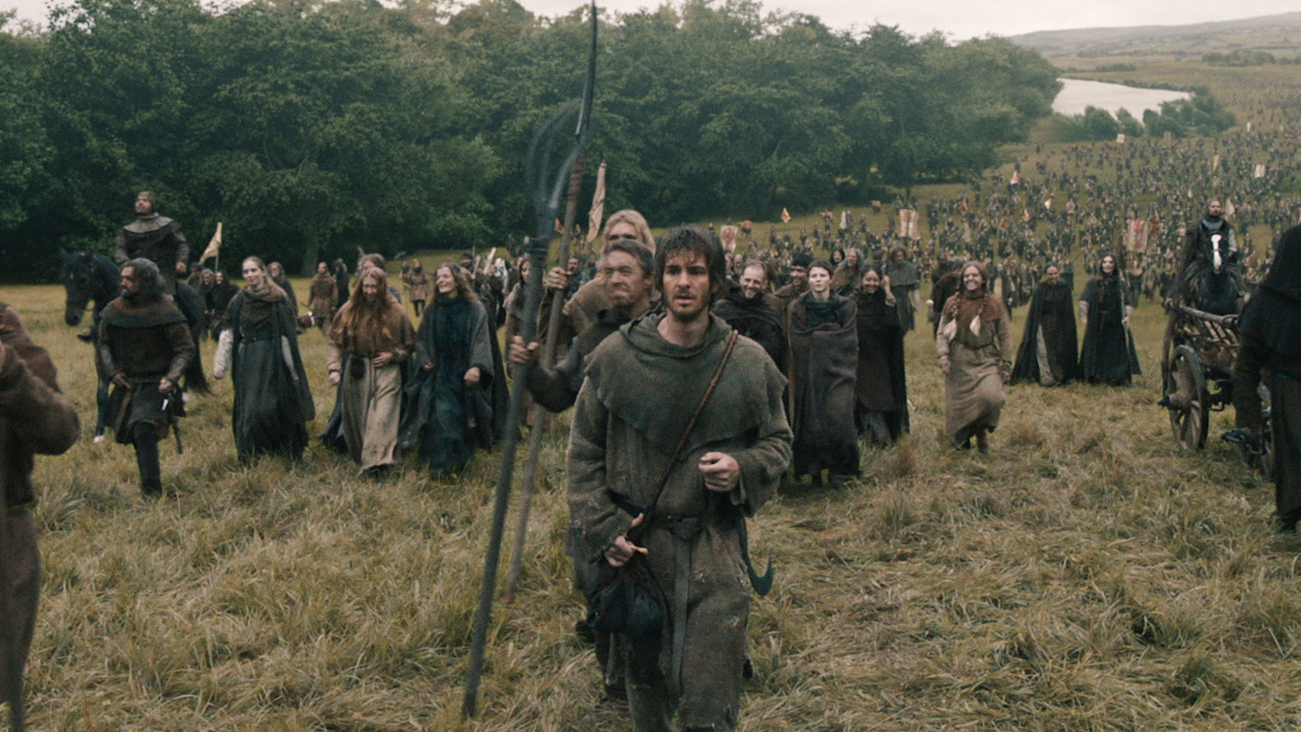 Andrew Garfield as Ploughman, Jamie Bell as John Ball, and Thomasin McKenzie as Alyce lead a large crowd of medieval peasants marching across a field.