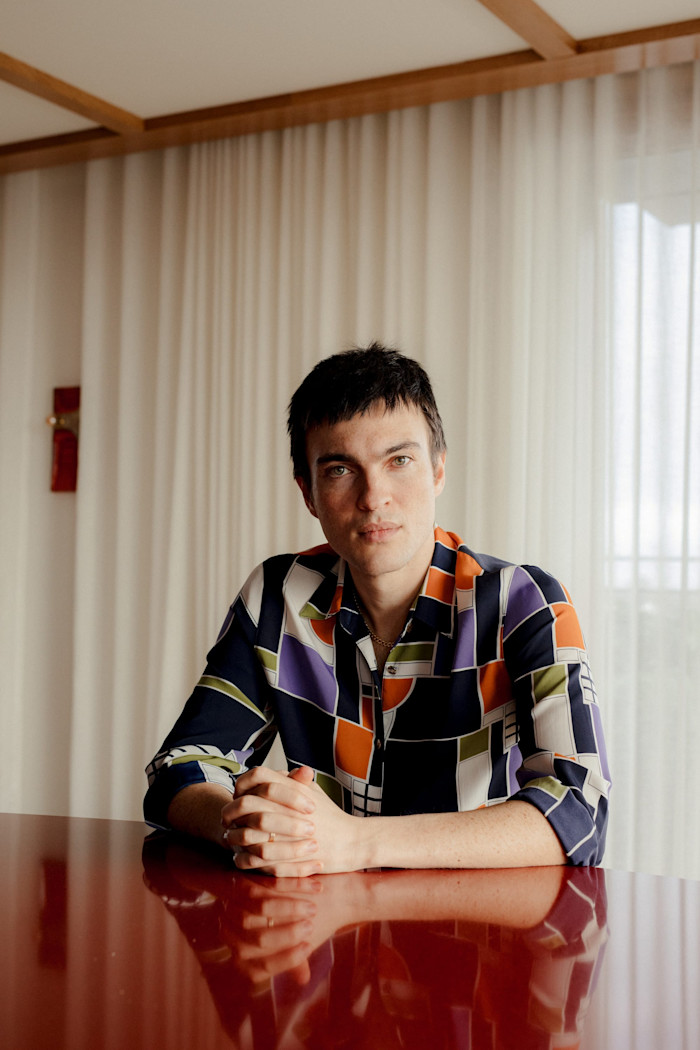 Mazdak Sanii sits at a red table with hands clasped, wearing a patterned shirt, in front of sheer curtains.