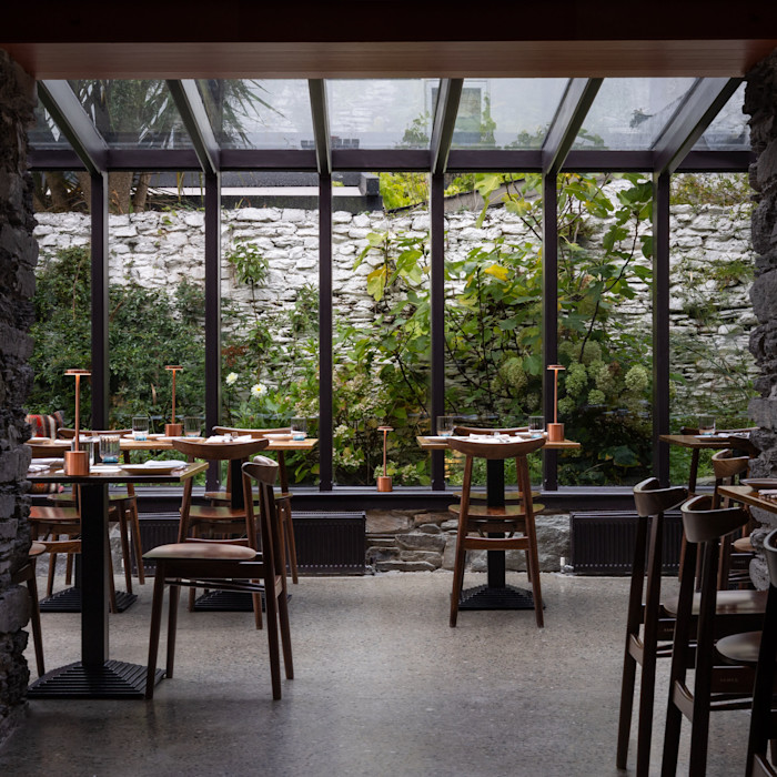 A dining area with wooden tables and chairs, set beside large glass windows overlooking a garden with stone walls and lush greenery.