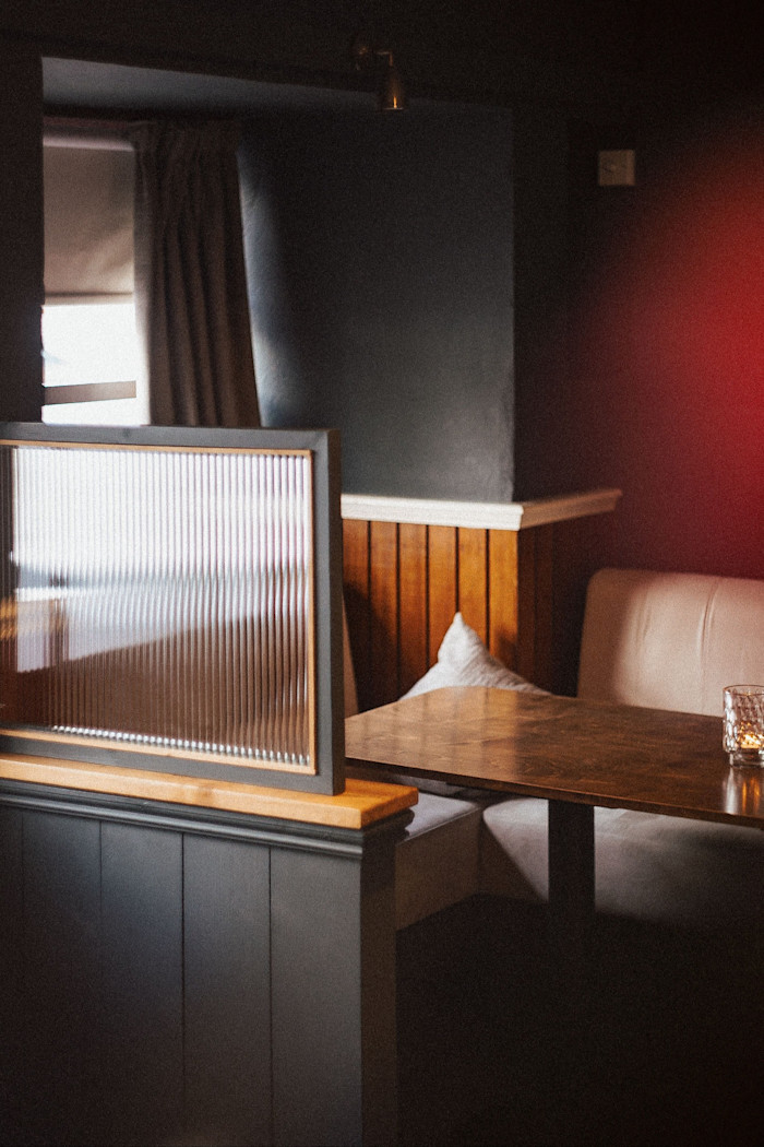 A wooden restaurant booth with beige cushioned seating, a dark wood table, a glass candle holder, and a ribbed glass partition.