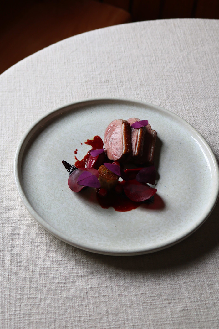 Slices of seared duck breast with ruby-red beetroot, sauce, and garnished with purple microgreens on a ceramic plate.