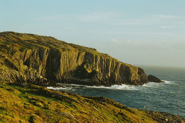 Steep rocky cliffs rise above the Atlantic Ocean, with waves crashing at their base and grassy slopes in the foreground.