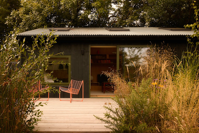 A small dark wooden cabin with green-trimmed windows and sliding doors, surrounded by tall grasses and shrubs. Two orange wire chairs sit on a wooden deck.