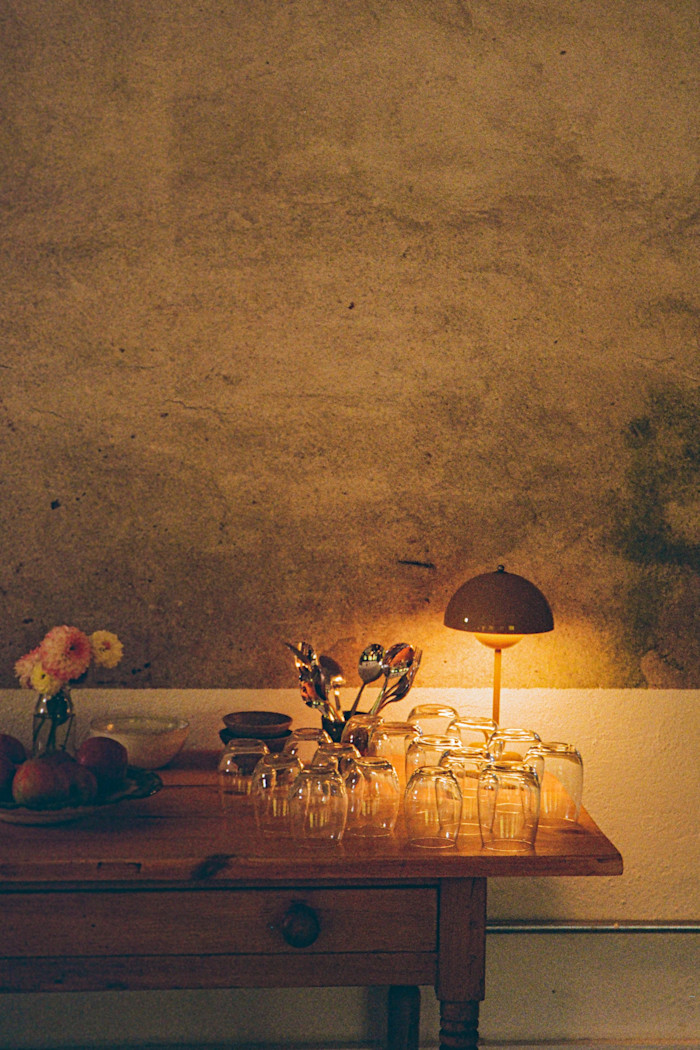 A wooden table set with a small lamp, glass tumblers, a bowl of fruit, a vase of flowers, and utensils, against a bare, textured wall.