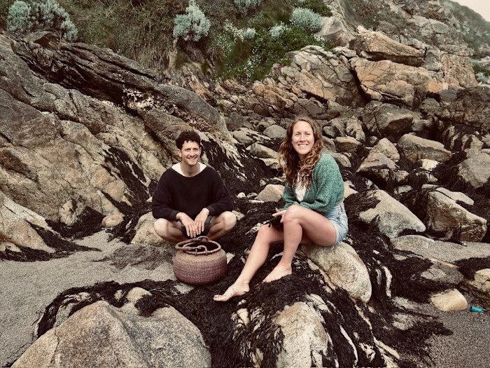 A man and a woman with a basket sitting on a rocky shoreline covered in seaweed.