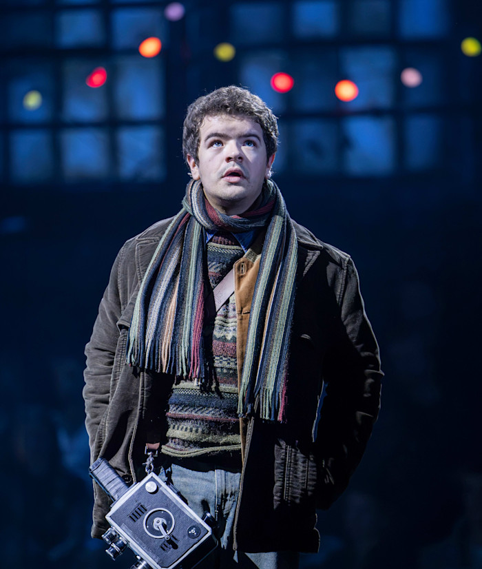 Gaten Matarazzo as Mark stands on stage in winter clothing and a striped scarf, holding a vintage camera, with colorful lights behind him.