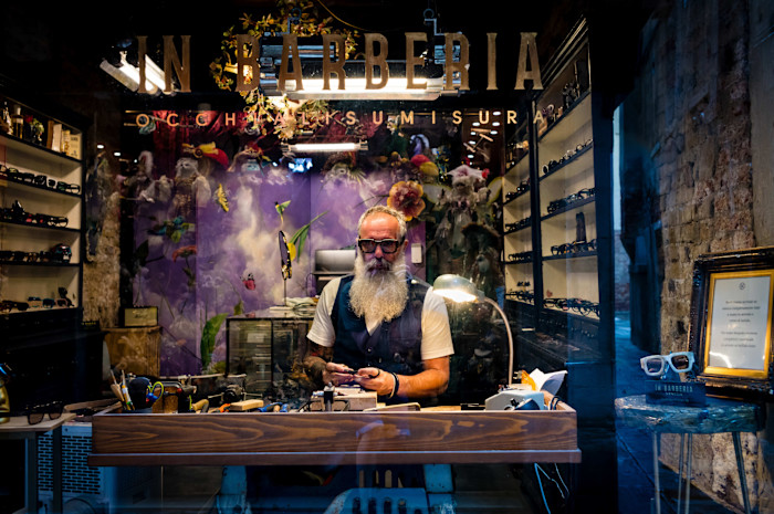 Antonio Battaglia, with silver hair, very long silver beard and tattoos, stands behind a workbench, with shelves of glasses around him.