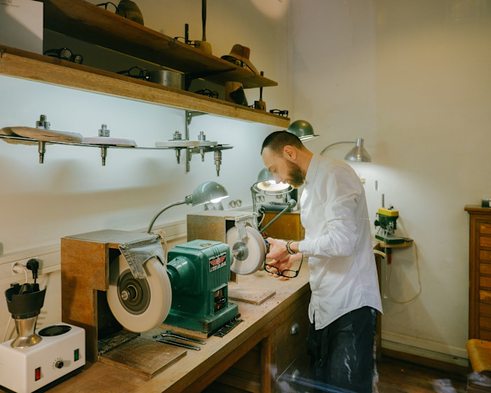 A young man uses a polishing machine at a workbench in a workshop filled with tools and lamps.