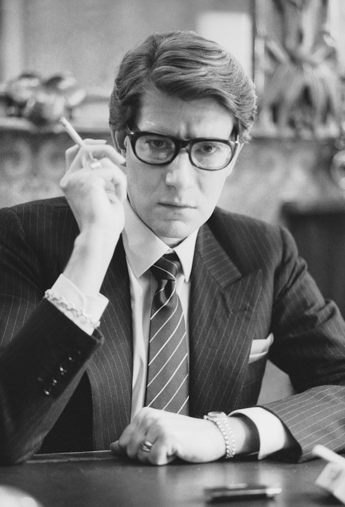 Yves Saint Laurent sits at a desk in his Paris studio, wearing a pinstripe suit, tie and glasses, and holding a cigarette.