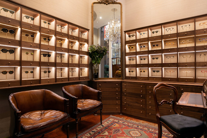 A desk, table and two leather armchairs in a room lined with cabinets where glasses are displayed.
