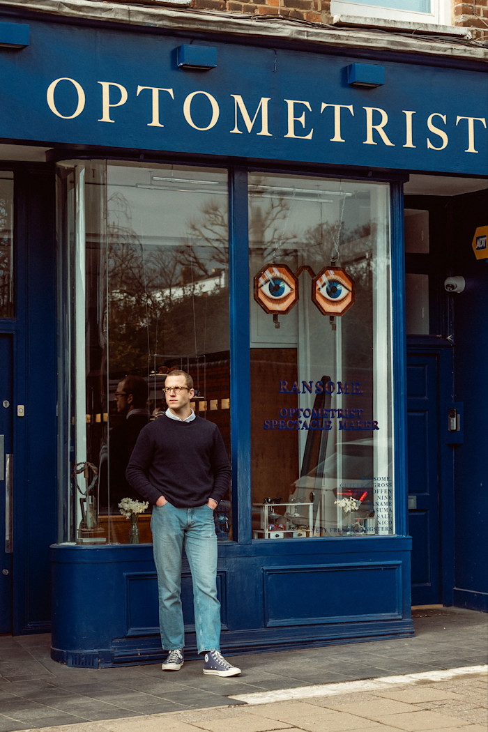 Jack Ransome Lupton, wearing jeans and a sweater, stands, hands in pockets, in front of his store.