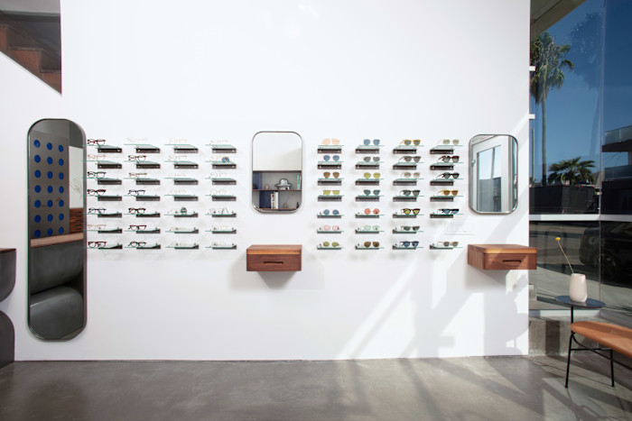 Rows of eyeglasses and sunglasses displayed on glass shelves against a white wall, with two mirrors and two small wooden drawers beneath them.