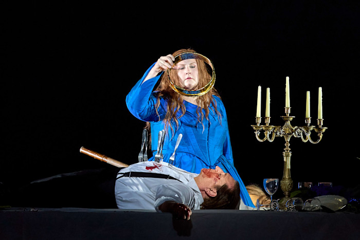 A woman in a blue dress holds a golden ring above a man lying on a table with blood and utensils stuck in his torso, next to a candelabrum.