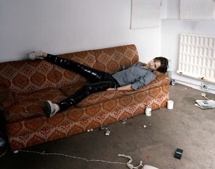 A young person reclines on a patterned orange sofa in a sparsely furnished, messy room, looking directly at the camera.