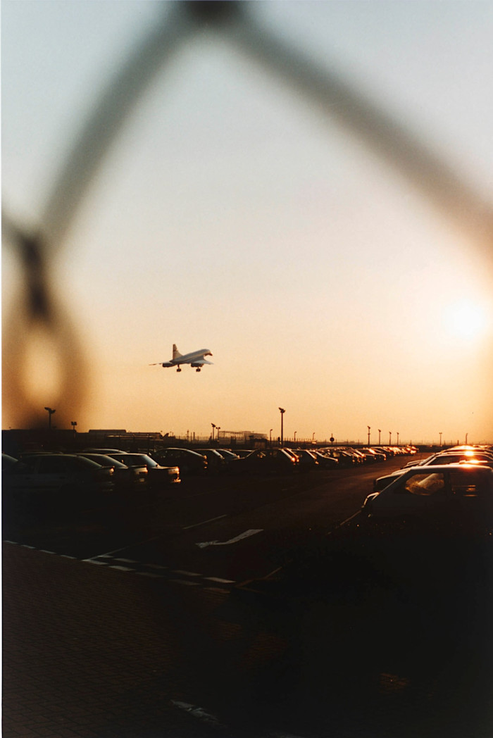 A Concorde jet approaches for landing over a parking lot at sunset, framed by an out-of-focus fence in the foreground.