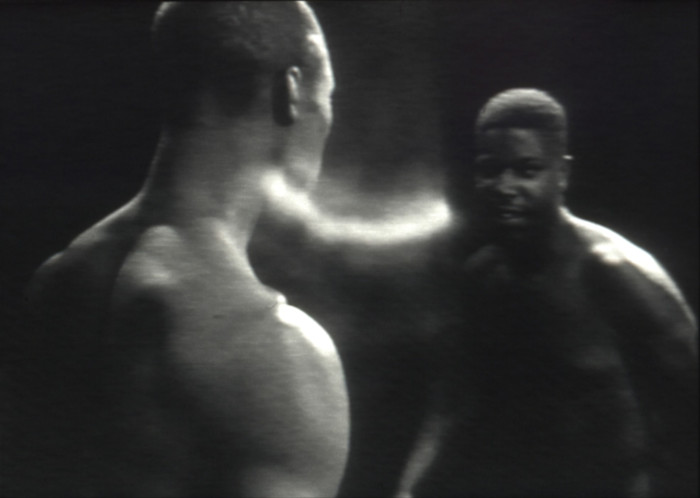 Two shirtless men stand facing each other in a dimly lit black-and-white scene.