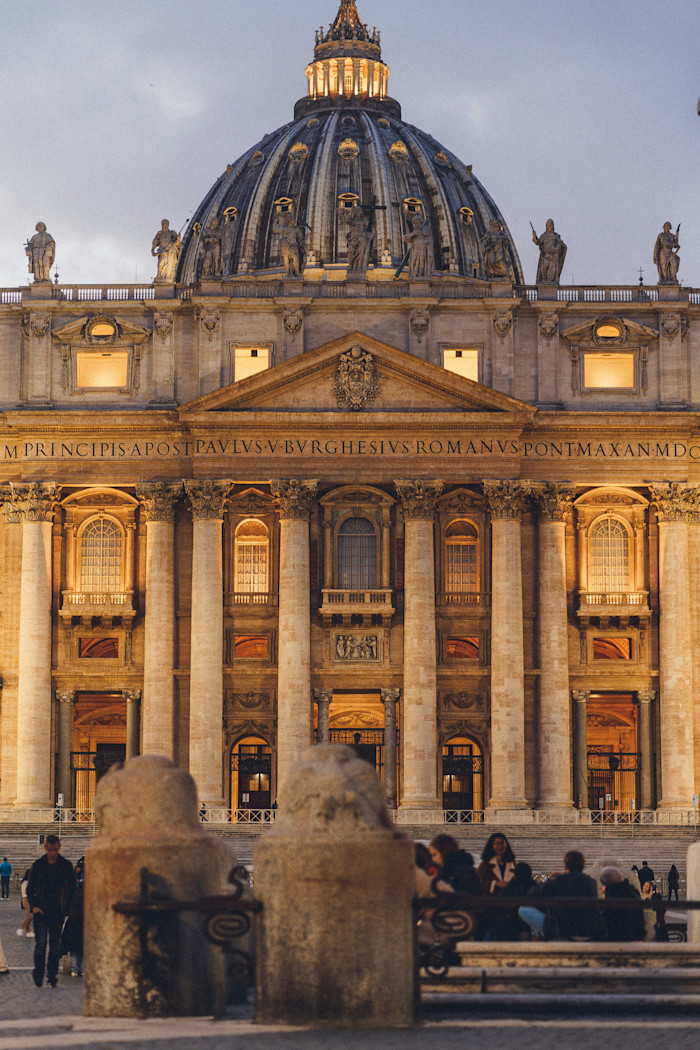 St. Peter's Basilica with its illuminated dome and facade, seen from the square with people gathered in the foreground.