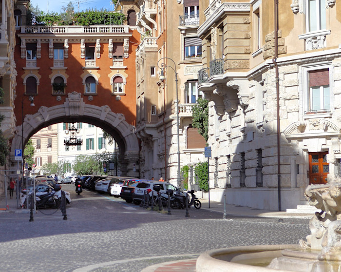 Ornate Art Nouveau arch with detailed facade connects two buildings in Rome’s Coppedè district, with parked cars and a fountain in view.