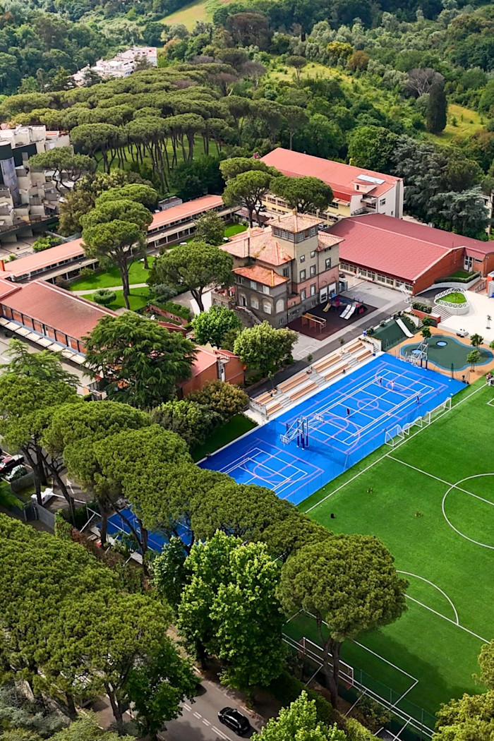 Aerial view of the American Overseas School of Rome campus, showing red-roofed buildings, a blue multi-sport court and a green soccer field surrounded by trees.