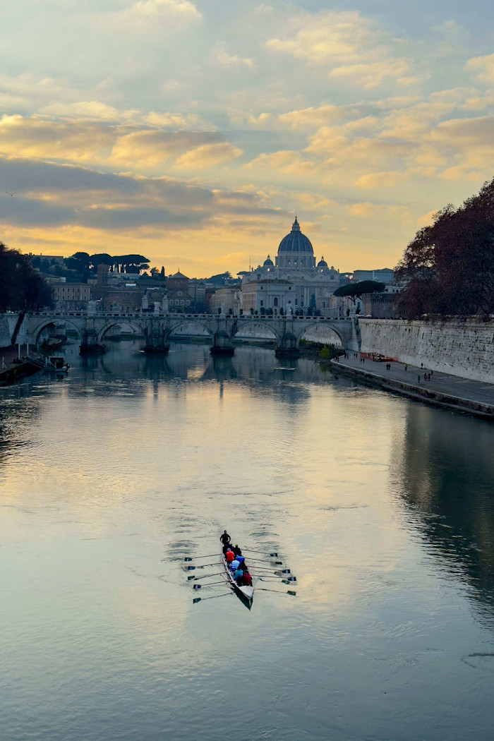 St. Peter's Basilica and Vatican City at sunset with a rowing team gliding along the Tiber River in the foreground.