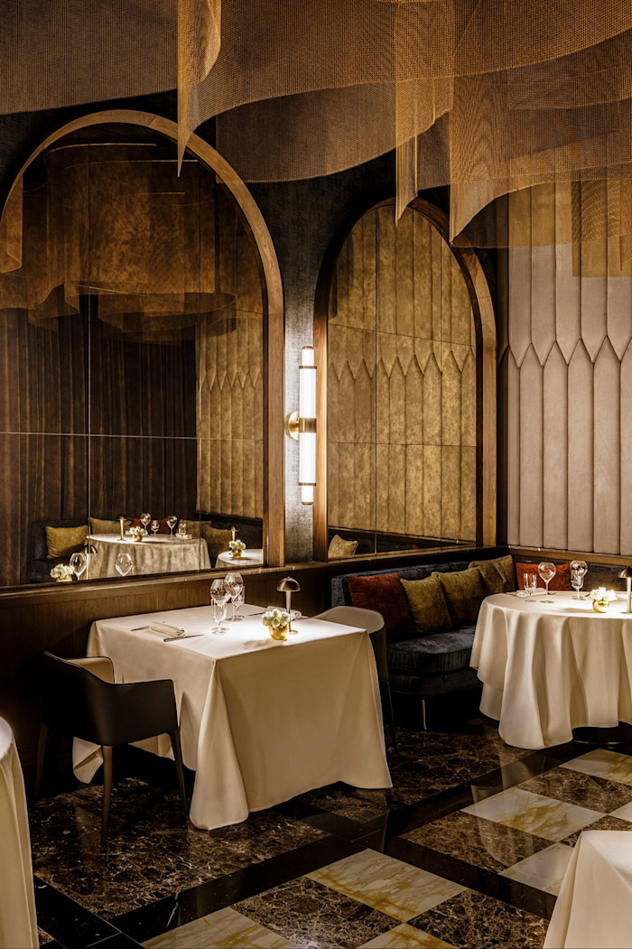 Elegant restaurant interior with tables covered in white linens, plush seating, gold decorative accents and dramatic lighting.