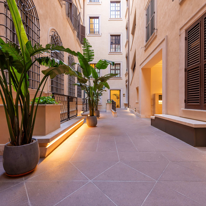 A courtyard with potted plants, warm lighting and beige stone walls lined with windows and shutters.