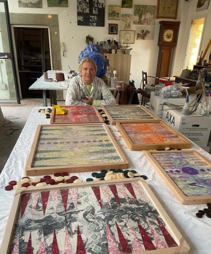 Artist seated in a cluttered studio behind a table covered with colourful patterned artworks and small game pieces.