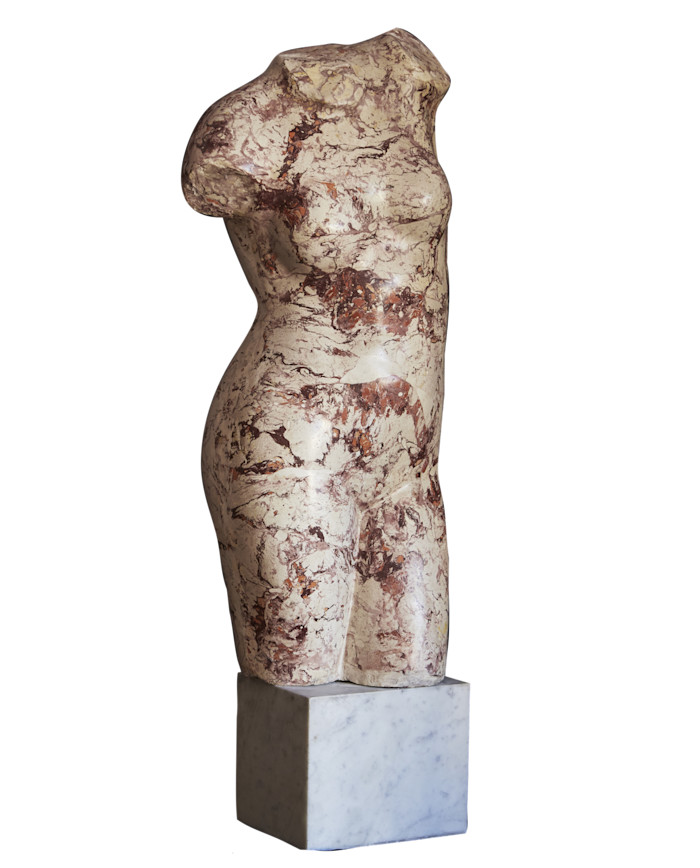 Headless female torso sculpture in mottled cream and reddish stone, displayed on a white marble plinth.