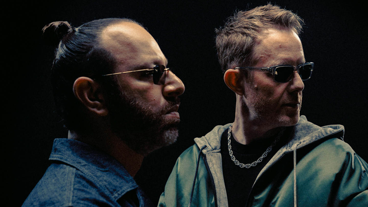 Two men wearing sunglasses in a dark setting, one with a top knot and beard, the other with short hair and a chain necklace.