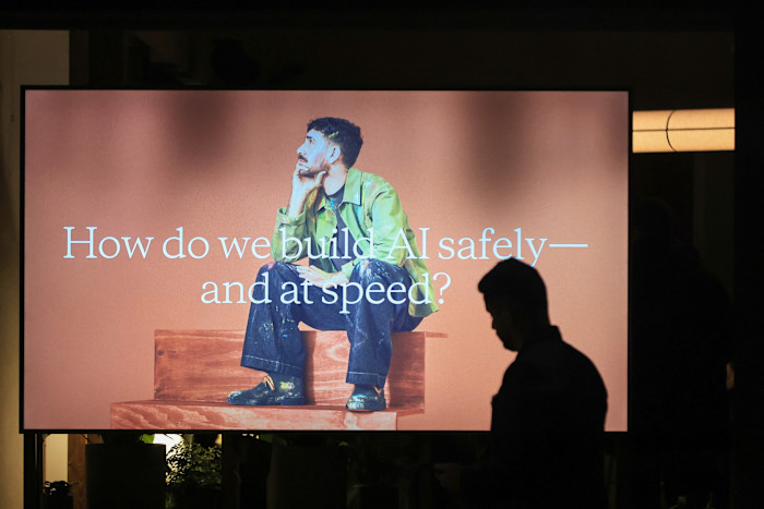 A person’s silhouette in front of a large screen displaying an image of a man sitting thoughtfully with the text, “How do we build AI safely—and at speed?”