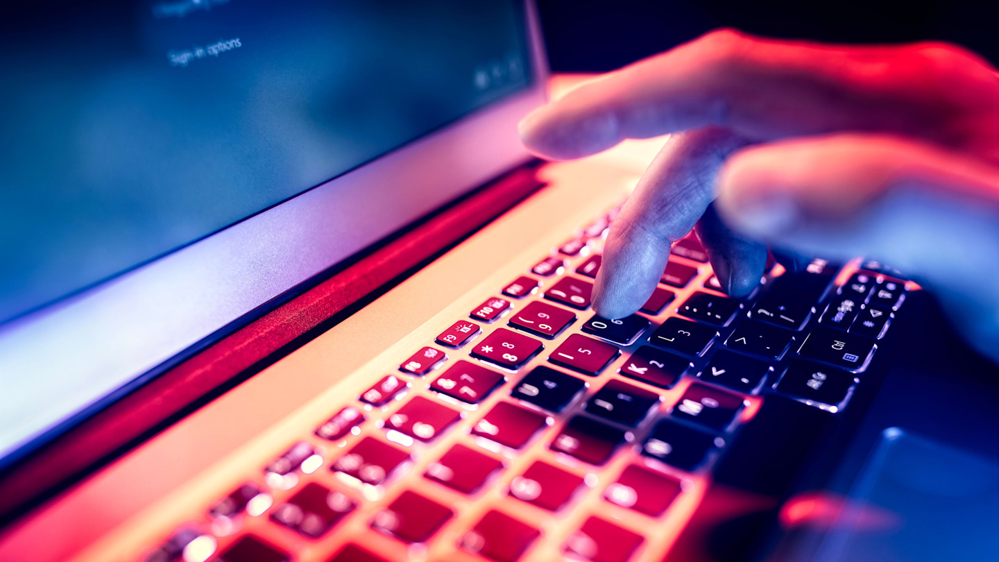 A hand typing on a laptop keyboard illuminated by the screen's glow in a dark environment.