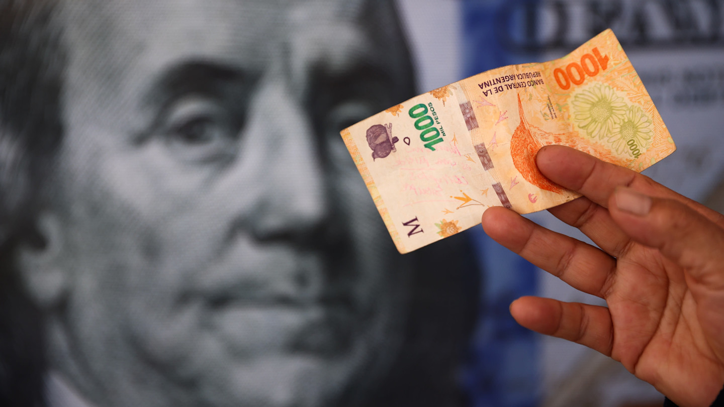 A hand holds a 1,000 Argentine peso banknote in front of a large, blurred image of a US dollar bill.