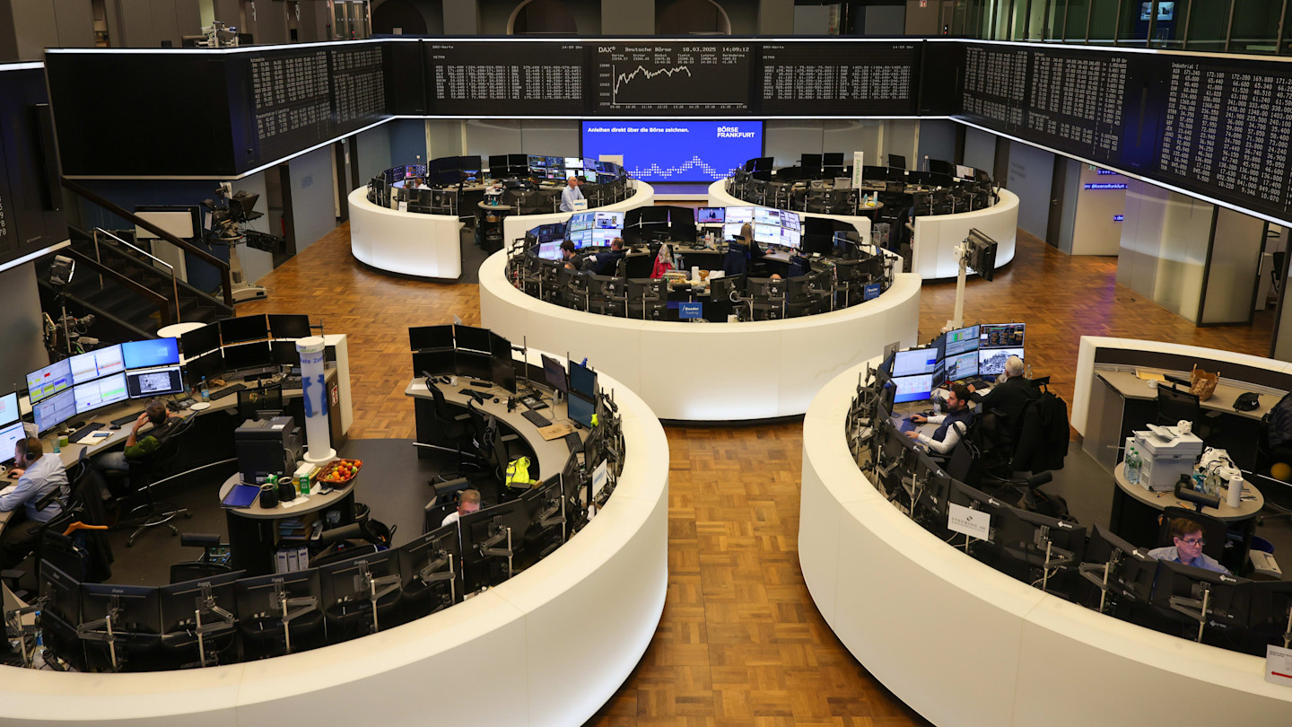 Traders work at computer stations inside the circular trading floor of the Frankfurt Stock Exchange, with DAX Index data displayed on large screens above.
