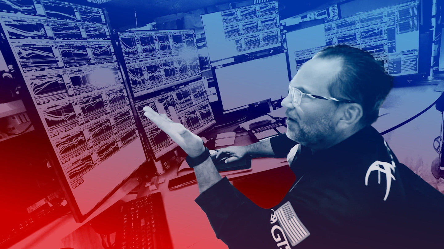 A trader gestures at a wall of computer monitors displaying financial charts and data in a trading room with a red and blue color overlay.
