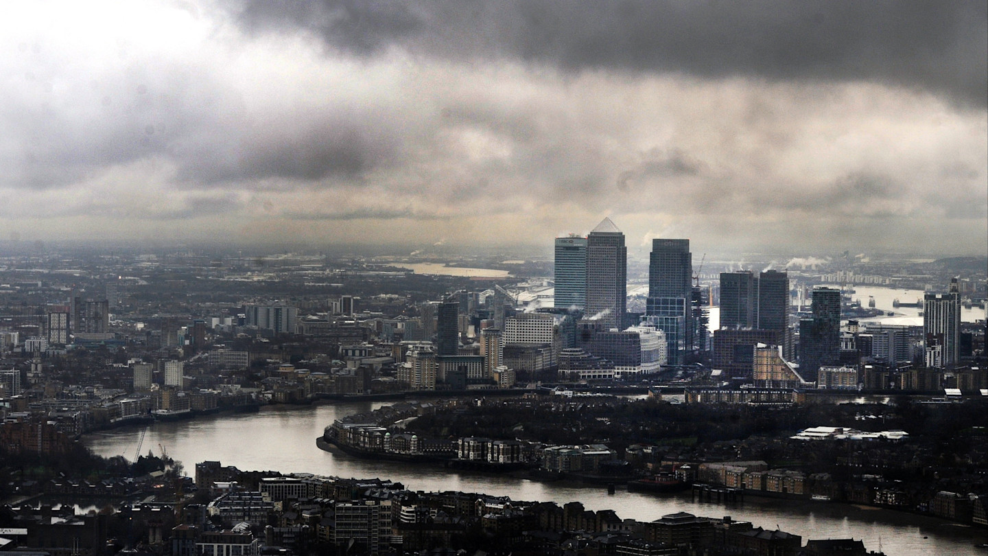 Dark clouds hang over the skyscrapers of London's Canary Wharf financial district, with the River Thames in the foreground.