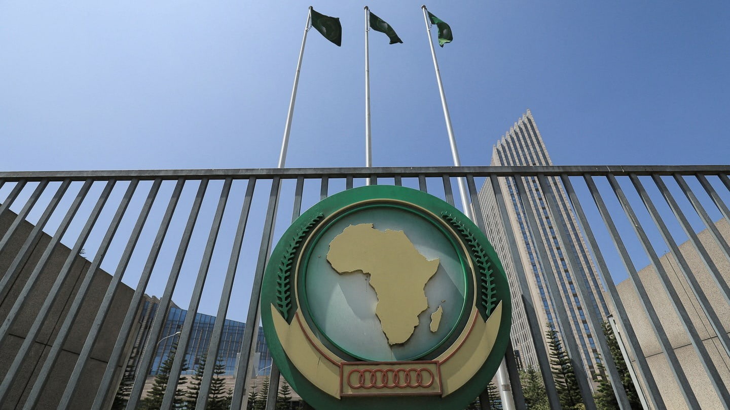 The African Union logo is displayed on a gate outside the AU headquarters building, with flagpoles and a high-rise in the background.