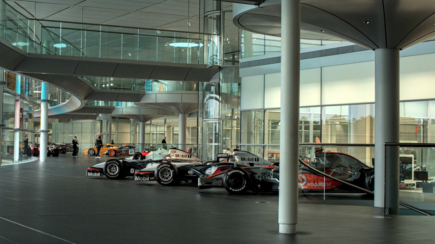 Several McLaren Formula 1 cars and other race cars are displayed in a modern glass-walled hall at the McLaren Technology Center.