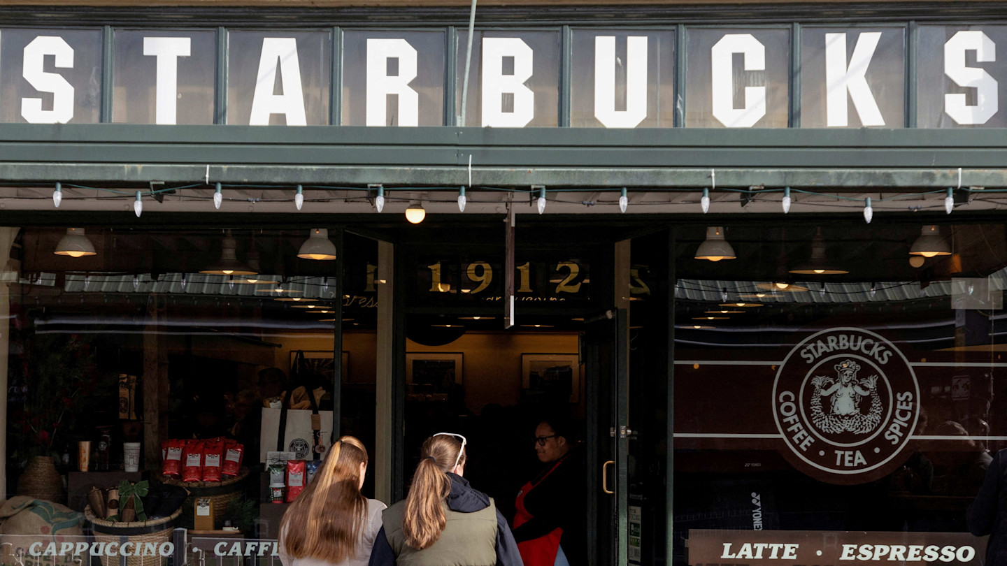 Customers wait outside the entrance of one of the first Starbucks stores at Pike Place Market, with the original Starbucks logo visible in the window.